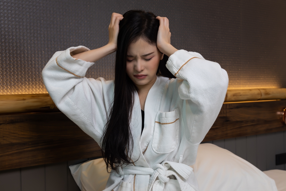 Young woman in bed with eyes shut holding the sides of her head in pain